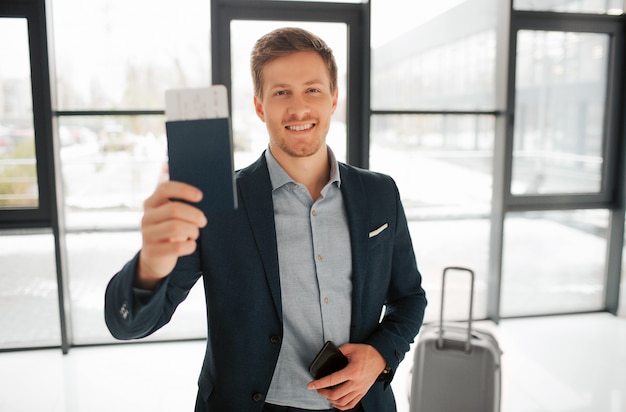 Feliz jovem buisnessman ficar no hall do aeroporto e mostrar o passaporte com bilhete