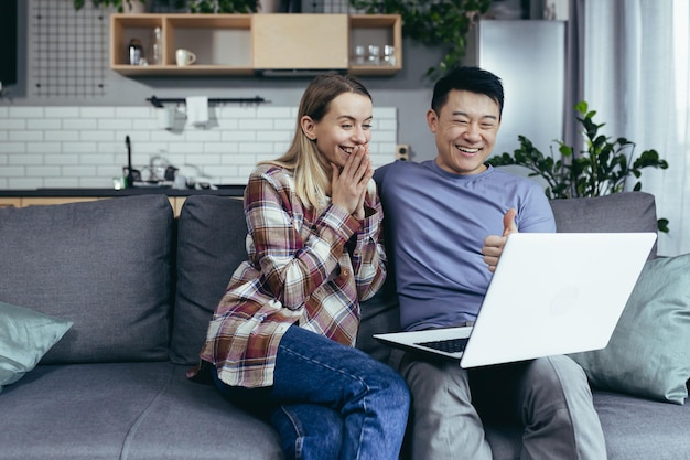 Feliz jovem casal asiático se unindo rindo usando laptop sentado à mesa em casa