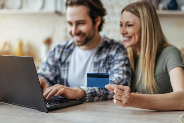 Feliz jovem casal caucasiano fazendo compras on-line e mostrando cartão de crédito no interior da cozinha turva