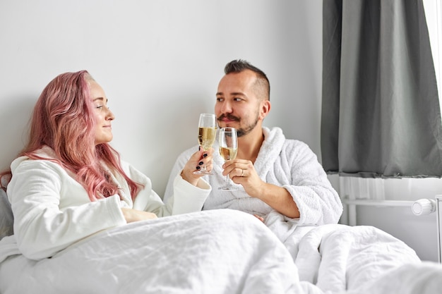 Feliz jovem casal caucasiano relaxando na cama, bebendo champanhe espumante em taças. a comemorar. fim de semana descanse relaxe