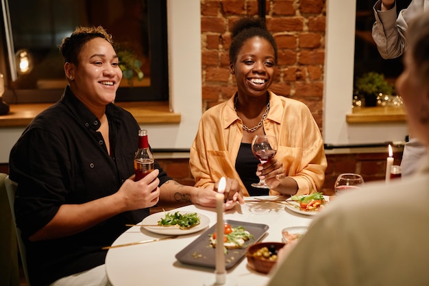 Feliz jovem casal de lésbicas com bebidas alcoólicas sentado à mesa servida e conversando com amigos que