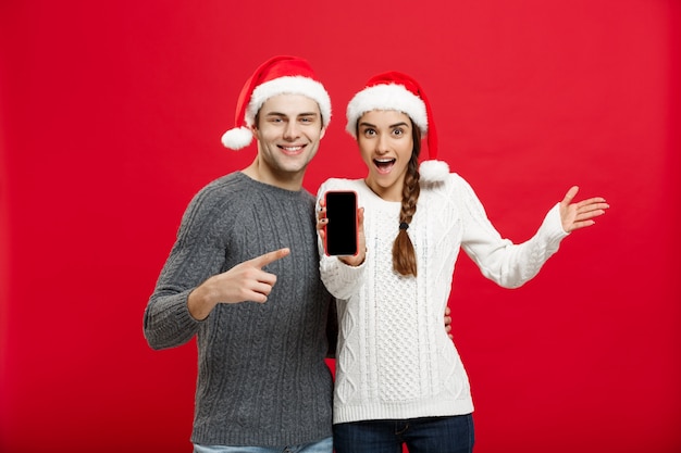 Feliz jovem casal em blusas de natal apontando o dedo com o telefone móvel.