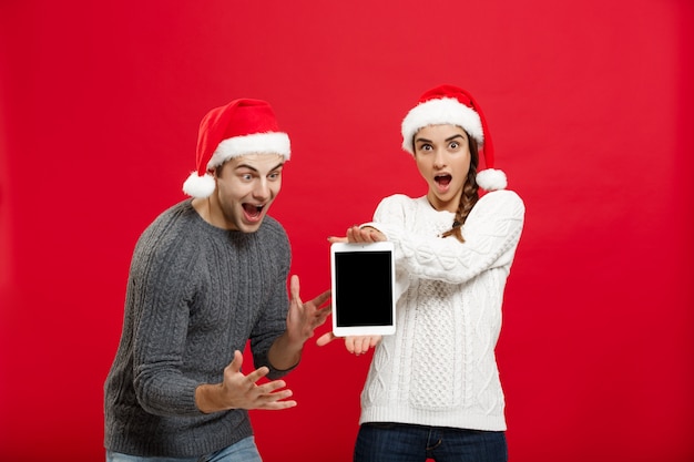 Feliz jovem casal em camisolas de Natal surpreende algo no tablet digital.