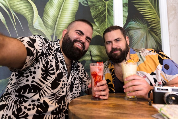 Feliz jovem casal gay em roupas de verão tirando um auto-retrato juntos enquanto toma um coquetel em suas férias