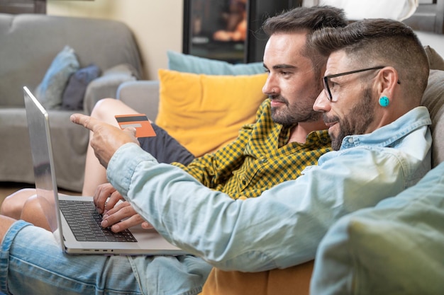 Feliz jovem casal gay usando laptop enquanto está sentado em um sofá em casa fazendo compras on-line com cartão de crédito