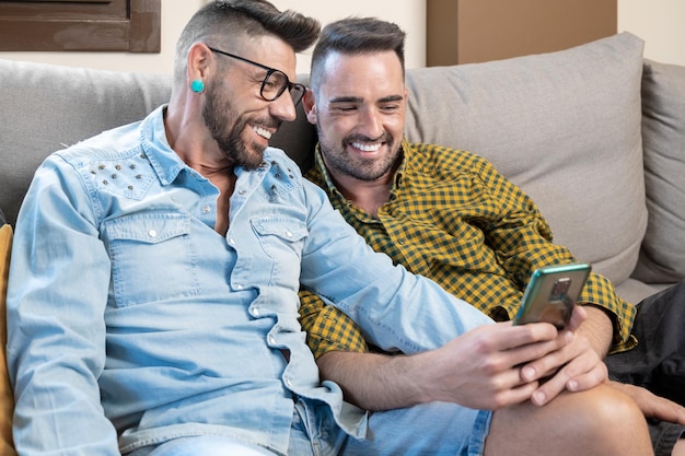 Feliz jovem casal gay usando telefone celular enquanto está sentado em um sofá na sala de estar