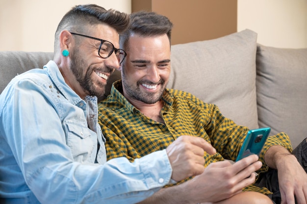 Feliz jovem casal gay usando telefone celular enquanto está sentado em um sofá na sala de estar