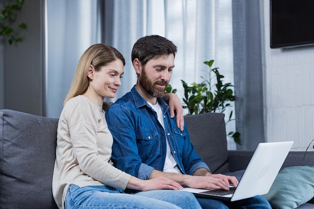 Feliz jovem casal homem e mulher juntos em casa sentados no sofá sorrindo e usando laptop