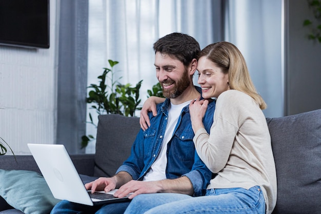 Feliz jovem casal homem e mulher juntos em casa sentados no sofá sorrindo e usando laptop