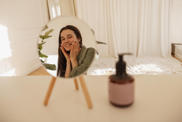 Feliz jovem caucasiana com pele fresca e saudável olhando no espelho enquanto está sentado à mesa morena sorri e toca seu rosto com as mãos enquanto aplica creme conceito de cuidados de beleza de mulher