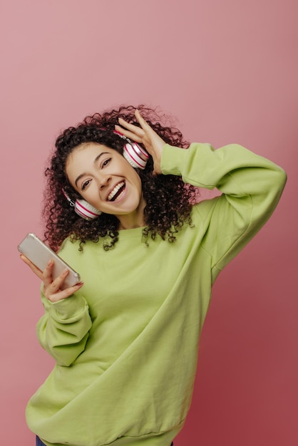 Feliz jovem caucasiana dançando e ouvindo música em fones de ouvido sem fio enquanto segura o smartphone no fundo rosa linda morena encaracolada usa blusa verde conceito de uso de telefone celular