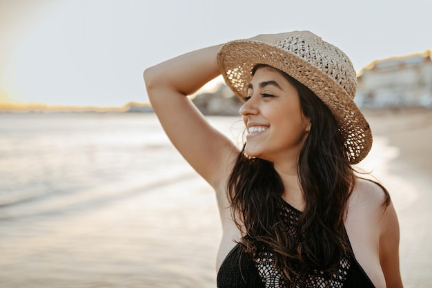 Foto feliz jovem caucasiana de maiô e chapéu de palha posando na praia olhando de lado e sorrindo