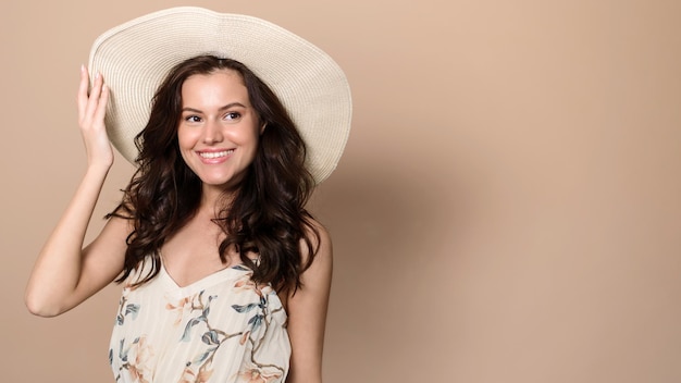 Feliz jovem caucasiana em vestido de verão e chapéu de palha olha para o espaço vazio no fundo do estúdio bege Senhora alegre anunciando seu produto de verão