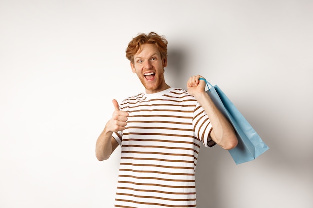 Feliz jovem com cabelo ruivo, fazendo compras nas lojas, mostrando o polegar para cima e segurando o saco de papel por cima do ombro, recomendando a loja, fundo branco.