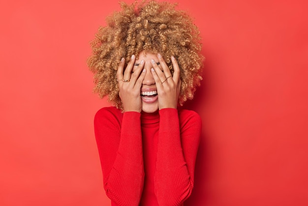 Feliz jovem de cabelos cacheados irreconhecível cobre os olhos com as mãos sorri abertamente se esconde de alguém usa gola rulê casual isolada sobre fundo vermelho vívido conceito de emoções humanas positivas