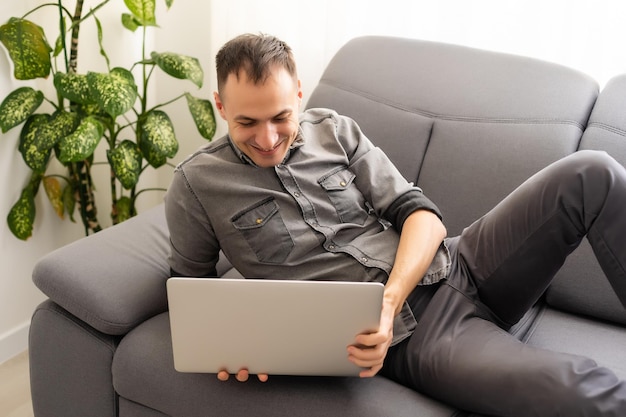 Feliz jovem de camisa no sofá em casa, trabalhando no computador portátil, sorrindo.