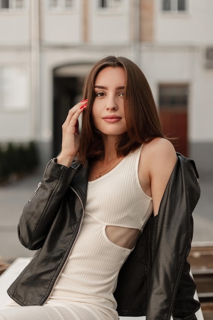 Feliz jovem e linda garota caucasiana em um vestido branco com uma jaqueta de couro da moda senta-se na rua e olha para a câmera