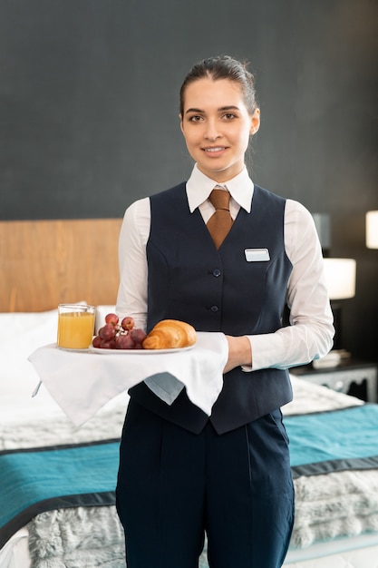 Foto feliz jovem elegante trabalhadora do hotel segurando a bandeja com café da manhã enquanto está em frente a câmera contra a cama e olhando para você