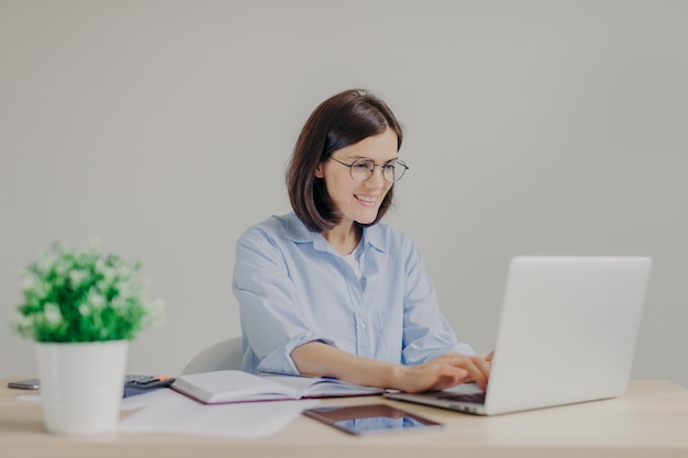 Feliz jovem empreendedor feminino em camisa casual e rodada grandes espetáculos analisa informações no computador portátil