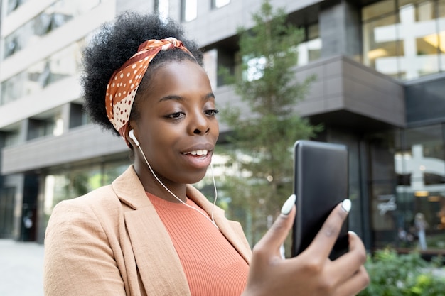 Feliz jovem empresária africana com fones de ouvido olhando para a tela do smartphone durante a comunicação em ambiente urbano