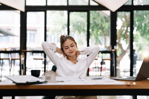 Feliz jovem empresária asiática relaxando com as mãos atrás da cabeça na mesa de escritório conceito de sonhar acordado