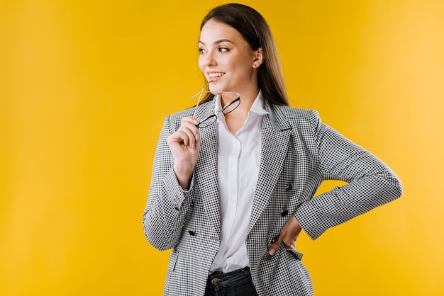 Feliz jovem empresária de casaco e camisa usando óculos sorrindo e posando para a câmera em fundo amarelo