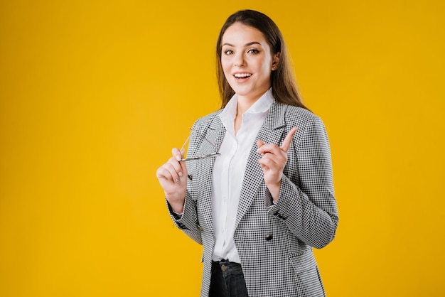 Feliz jovem empresária de casaco e camisa usando óculos sorrindo e posando para a câmera em fundo amarelo