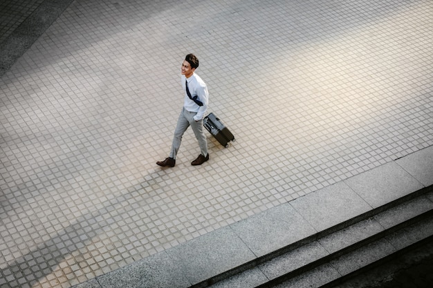 Feliz jovem empresário andando com mala na cidade. Macho moderno em viagem de negócios. Vista de alto ângulo. Comprimento total