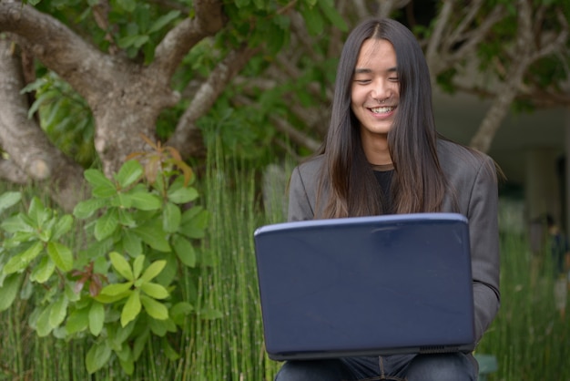 Feliz jovem empresário asiático com cabelo comprido, usando laptop no parque