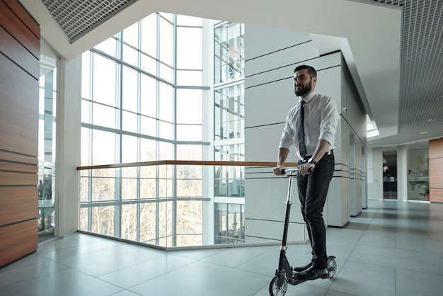 Feliz jovem empresário barbudo avançando em pé na scooter dentro do centro de negócios contemporâneo