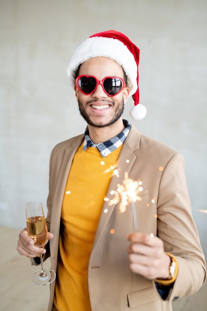 Foto feliz jovem empresário em roupas casuais, boné de papai noel e óculos de sol em forma de coração segurando uma taça de espumante champanhe e luz de bengala