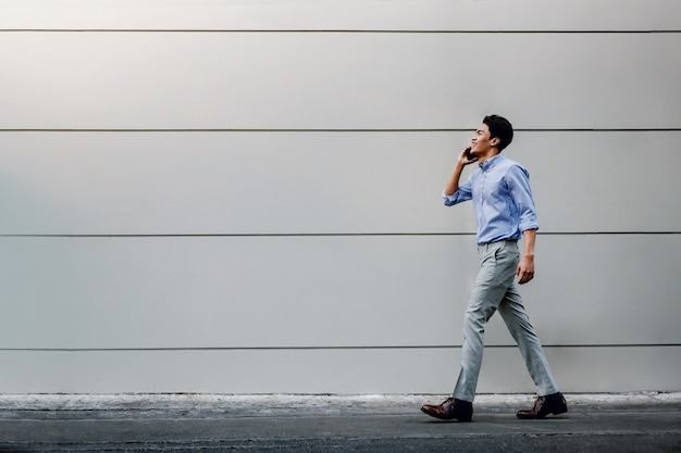 Feliz jovem empresário em roupas casuais, usando telefone celular enquanto caminhava pela parede do edifício urbano. Estilo de vida das pessoas modernas.