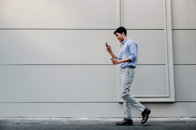 Feliz jovem empresário em roupas casuais, usando telefone celular enquanto caminhava pela parede do edifício urbano. Estilo de vida das pessoas modernas.
