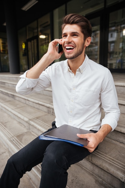 Feliz jovem empresário segurando uma pasta com documentos e falando no celular ao ar livre