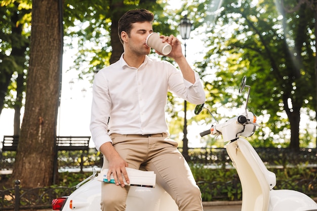 Feliz jovem empresário sentado em uma moto ao ar livre, lendo jornal, bebendo café