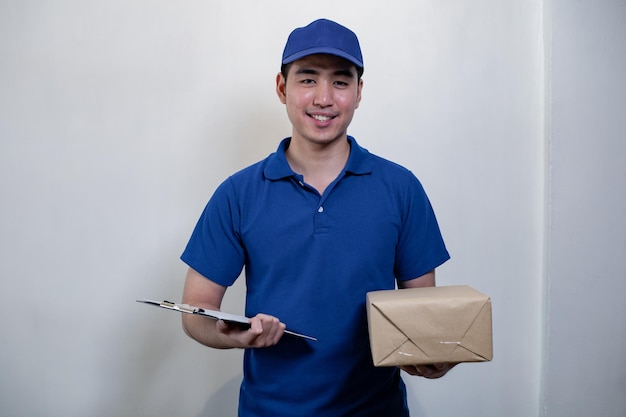 Feliz jovem entregador de vestido azul com caixa postal e documento sobre fundo branco com sorriso. sorrindo de homem jovem correio entrega postal masculino na entrega do pacote. conceito de entrega.