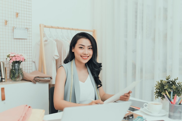 Feliz jovem estilista asiático em trajes casuais sentado à mesa e fazendo esboços de moda