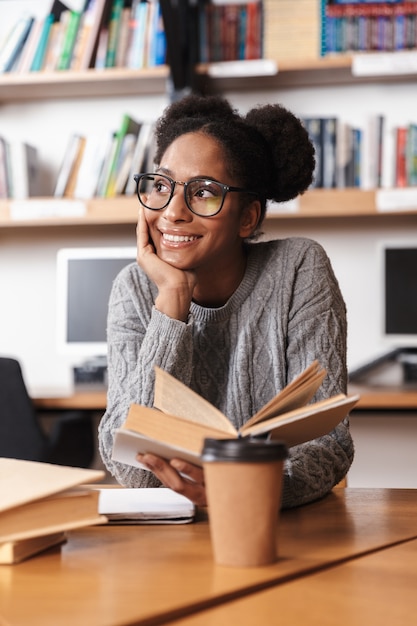 Feliz jovem estudante africana estudando na biblioteca, lendo um livro