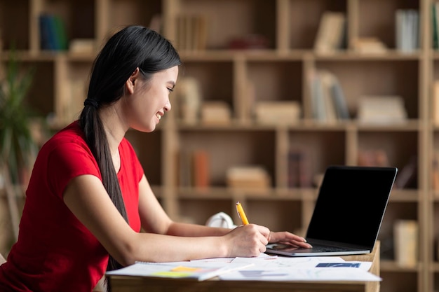 Feliz jovem estudante asiática assistindo aula on-line no laptop com tela vazia no gabinete