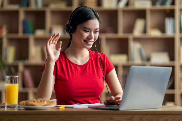 Foto feliz jovem estudante asiática em fones de ouvido estudando com laptop cumprimentando acenando com a mão assistindo aula on-line