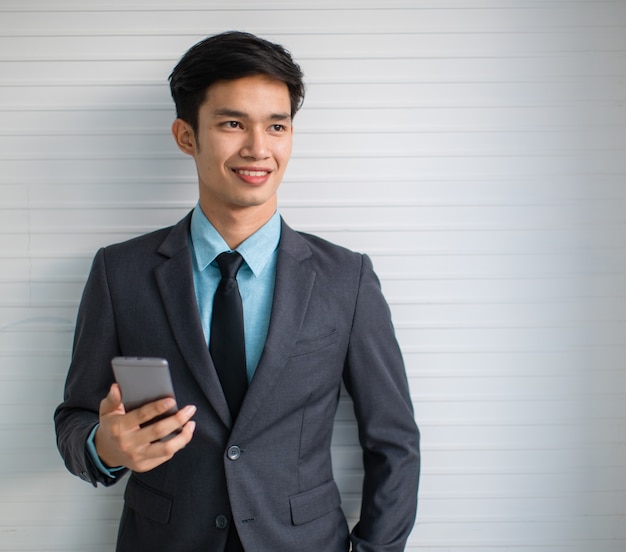 Feliz jovem gerente asiático de terno sorrindo e navegando no celular em pé contra a parede