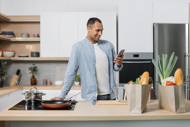 Feliz jovem indiano usando smartphone parado na cozinha pela manhã