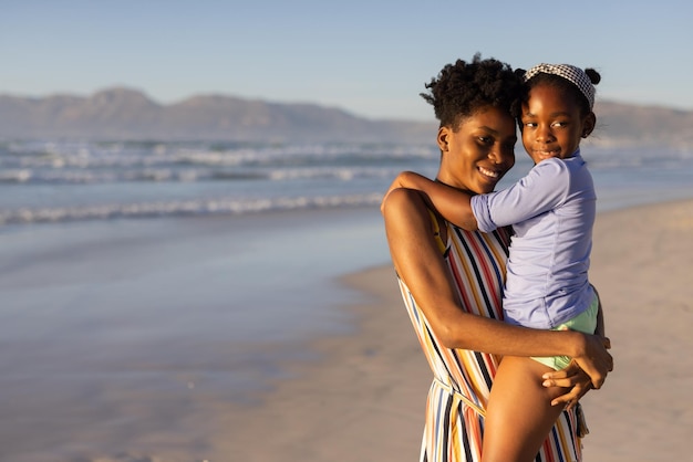 Feliz jovem mãe afro-americana carregando uma menina em pé contra o mar e o céu claro ao pôr do sol