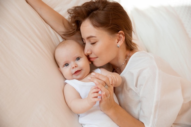 Foto feliz jovem mãe com bebê na sala branca mãe está deitada beijando bebê em casa na cama no quarto