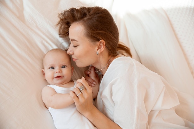 Feliz jovem mãe com bebê na sala branca mãe está deitada beijando bebê em casa na cama no quarto