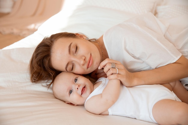 Feliz jovem mãe com bebê na sala branca mãe está deitada beijando bebê em casa na cama no quarto