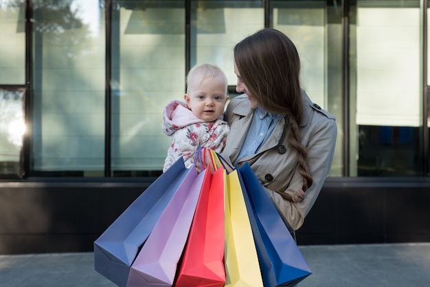 Feliz jovem mãe com filha nos braços e sacos de compras na mão.