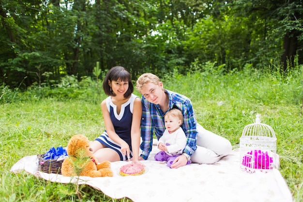 Foto feliz jovem mãe e pai com sua filha bebê relaxando em um cobertor em um parque, comemorando com bolo de aniversário.