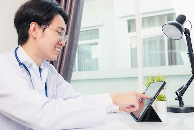 Feliz jovem médico asiático, homem bonito, sorriso, trabalho de escritório em casa, use óculos, usando um tablet digital moderno e preto preto na mesa do escritório do hospital, conceito de saúde e medicina de tecnologia
