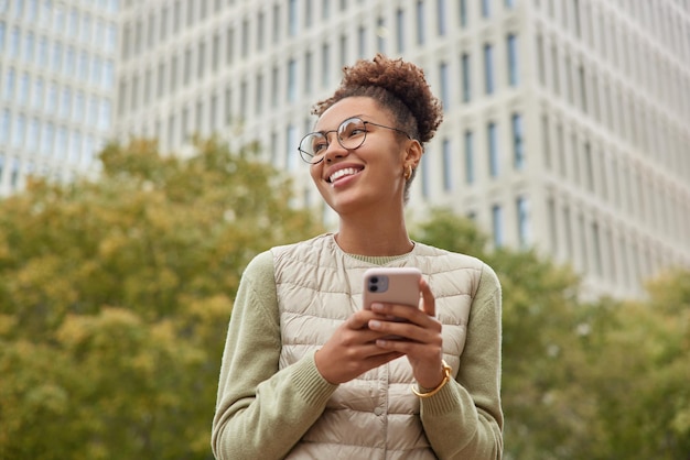 Foto feliz jovem milenar com cabelo encaracolado olhando para o outro lado fazendo passeios de celular na cidade enviando mensagens de texto usando roupas casuais